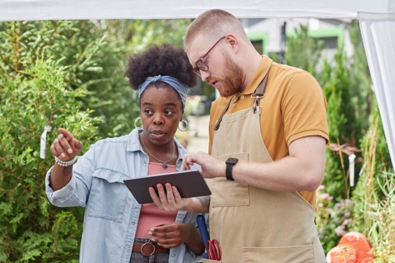 Medium shot of African American woman landscape designer pointing at exact coniferous shrub choosing plants for garden design, while talking to male horticulturalist using digital tablet outdoors.